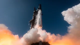 Rocket blasting off from a launch pad, with bright flame and white smoke at the base against a clear blue sky.