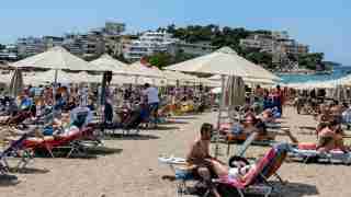 Crowded beach scene with rows of sun loungers and white umbrellas, people sunbathing and swimming in the blue sea.