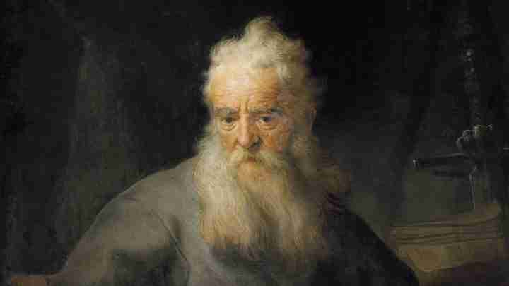 Portrait of an elderly man with a long white beard and solemn expression against a dark background, in a muted robe.