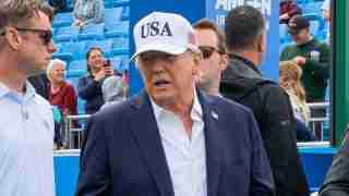 Man in a navy blazer and white shirt wearing a USA cap at an outdoor event with blue stadium seating behind him.
