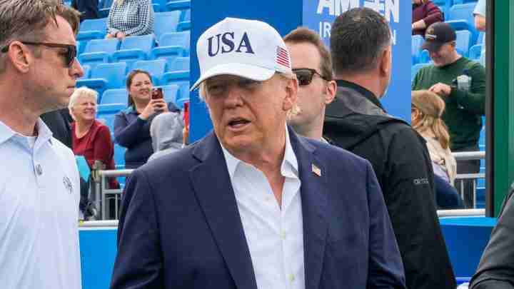 Man in a navy blazer and white shirt wearing a USA cap at an outdoor event with blue stadium seating behind him.