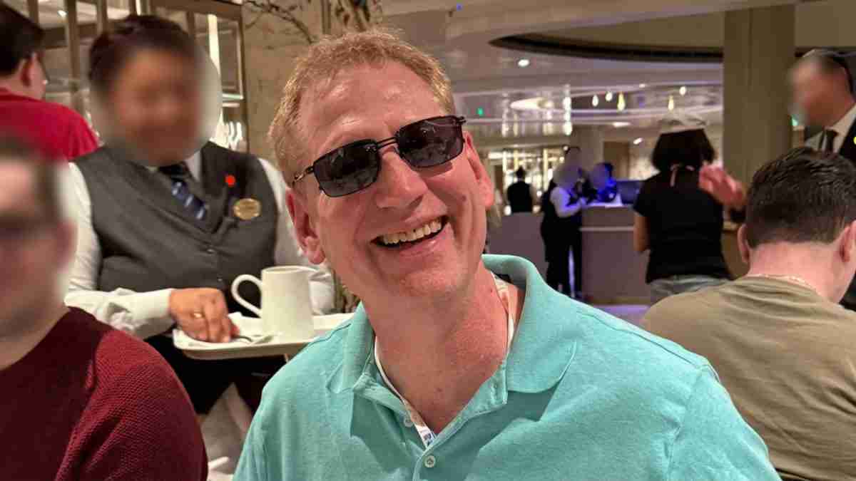 Smiling man with blond hair wearing sunglasses and a teal polo shirt, seated at a restaurant table with blurred patrons in the background.