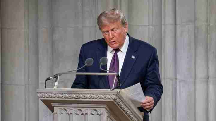 Donald Trump in a dark suit and purple tie speaks at a stone lectern with two microphones, holding papers.
