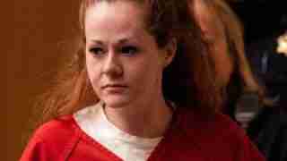Woman with long reddish-brown hair in a red jumpsuit, looking to the left in an indoor setting with a plain background on the left.