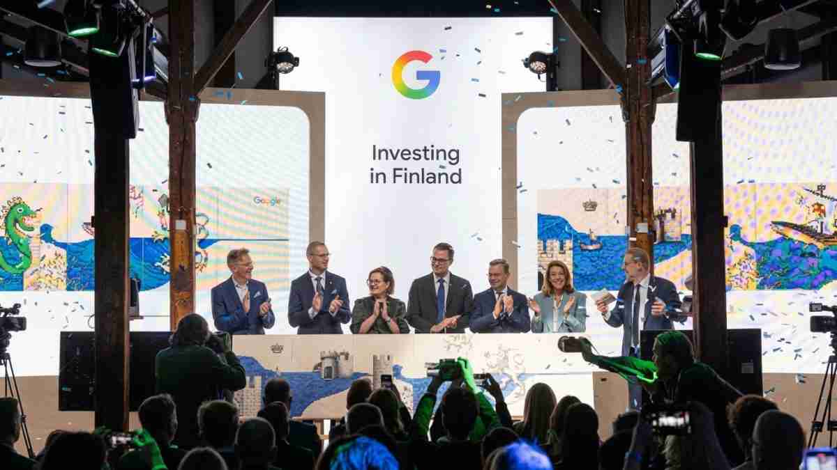 Group of eight people on a stage applauding at a conference, with a large screen reading 'Investing in Finland' and a Google logo above; confetti in the air; audience and cameras in foreground.