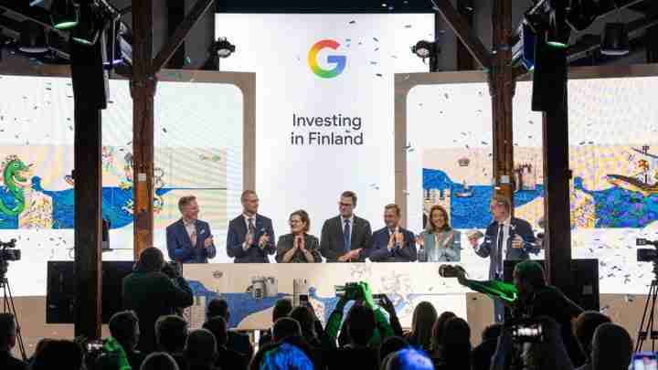 Group of eight people on a stage applauding at a conference, with a large screen reading 'Investing in Finland' and a Google logo above; confetti in the air; audience and cameras in foreground.